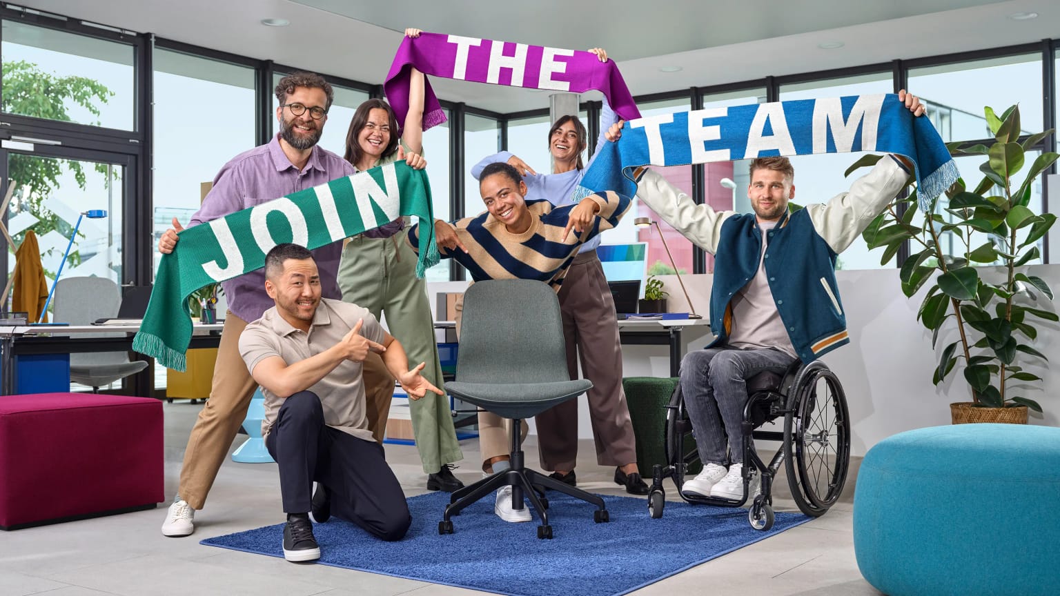 Sechs Personen in einem modernen Büro halten „JOIN THE TEAM“-Schals. Eine Person sitzt im Rollstuhl, eine andere zeigt auf einen freien Stuhl in der Mitte.